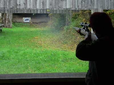 Eine Person zielt mit einem Gewehr auf eine entfernte Zielscheibe. Der Schießstand ist von Bäumen und Gras umgeben, die Umgebung wirkt herbstlich und bewaldet.